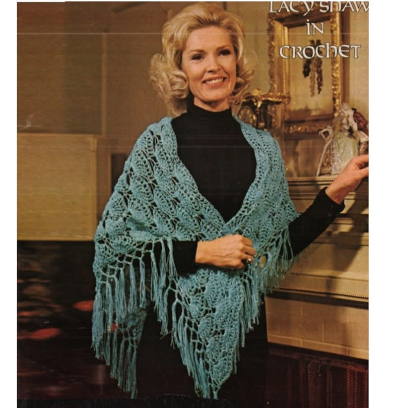 May include: A woman wearing a teal crochet shawl with fringe. The shawl has a lacy pattern and is draped over her shoulders. The text "Lacy Shaw in Crochet" is visible in the top right corner of the image.