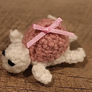Pode incluir: Uma tartaruga de crochê feita à mão com casco rosa e corpo branco. A tartaruga tem um pequeno laço rosa no casco e olhos pretos. Está sobre uma superfície de madeira.