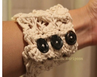 May include: A crocheted cuff with three black buttons. The cuff is made of a cream-colored yarn and has a textured design. The text "Lilou Mariposa" is visible on the cuff.