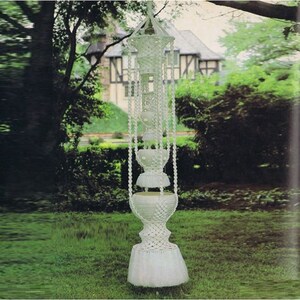May include: A white macrame hanging planter with three tiers. The planter is made of knotted rope and has a fringed bottom. It is hanging in a garden setting.