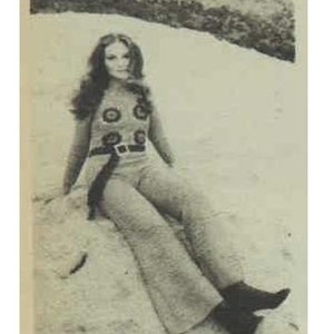 May include: A woman with long brown hair wearing a brown pantsuit with a black belt and a white and brown patterned top. She is sitting on a rock with her legs crossed.