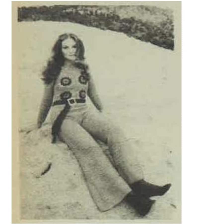 May include: A woman with long brown hair wearing a brown pantsuit with a black belt and a white and brown patterned top. She is sitting on a rock with her legs crossed.