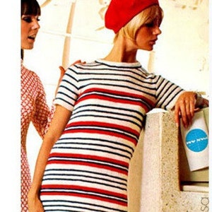 May include: A woman wearing a red beret and a white and red striped dress. The dress has a short sleeve and a fitted silhouette. The woman is standing next to a wall with a blue and white sign.