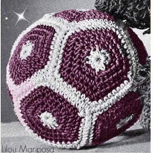 May include: A crocheted ball with a geometric pattern in shades of purple, white, and pink. The ball is made of yarn and has a textured surface.