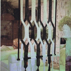 May include: A black and white macrame room divider with a wooden dowel rod. The divider is made of thick cords and has a diamond pattern. The divider is hanging in a room with a white couch and a white rug.