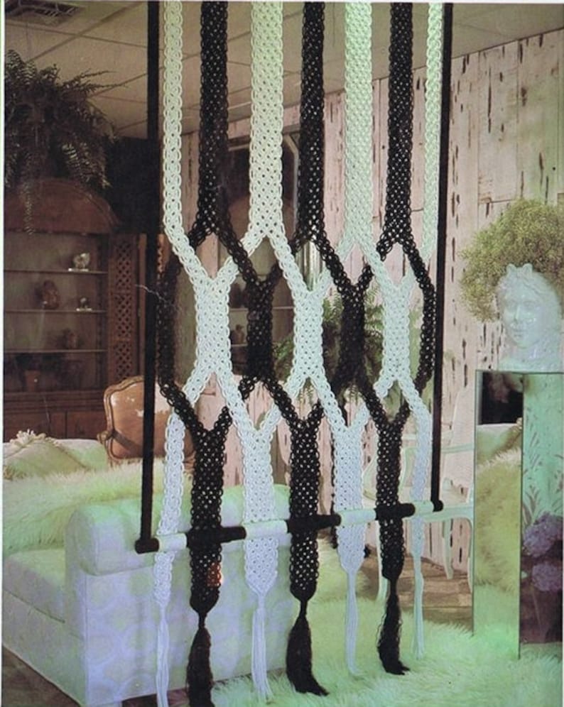 May include: A black and white macrame room divider with a wooden dowel rod. The divider is made of thick cords and has a diamond pattern. The divider is hanging in a room with a white couch and a white rug.