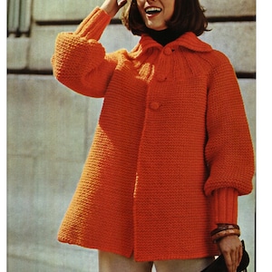 May include: A bright orange knitted cardigan with a button fastening and a wide collar. The cardigan has long sleeves with a slightly puffed look. The model is wearing the cardigan with a turtleneck and beige trousers.