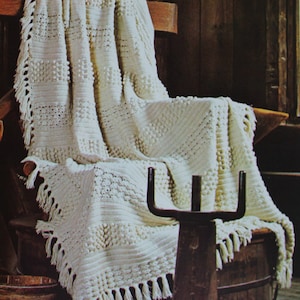 May include: A white crocheted blanket with a bobble stitch pattern and fringe trim draped over a wooden chair. The blanket is in a rustic setting with a wooden barrel and a metal stand.