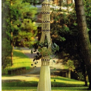 May include: A macrame plant hanger with a white cord and a brown pot with a green plant hanging from it. The hanger is in the shape of a tower with a bell-shaped top. The hanger is hanging in a garden setting.