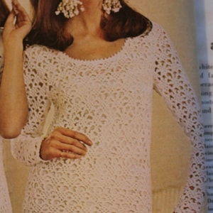 Puede incluir: Un vestido blanco de crochet con un delicado patrón de encaje. El vestido tiene mangas largas y un escote redondo.