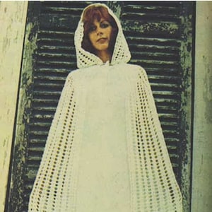 May include: A white crocheted hooded cape with a lacy pattern. The cape is worn by a woman with red hair and is photographed against a wooden door.