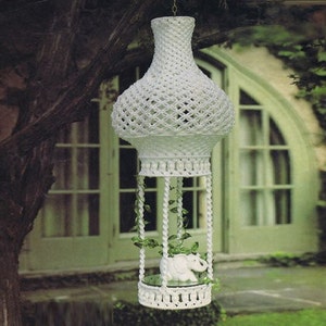 May include: White macrame plant hanger with a round base and a hanging planter. The planter is filled with a green plant and a small white elephant figurine.
