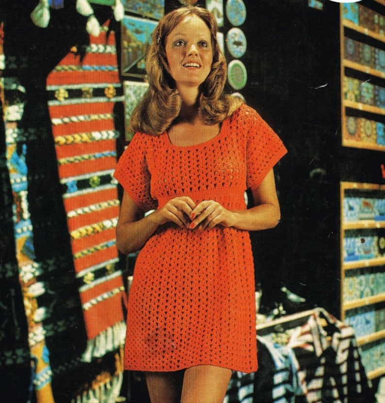 May include: A woman wearing a bright orange crochet mini dress with short sleeves. The dress has a loose fit and a crocheted pattern.