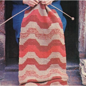 May include: A person is knitting a red and beige striped rug with a chevron pattern. The rug is being knitted on large wooden knitting needles.