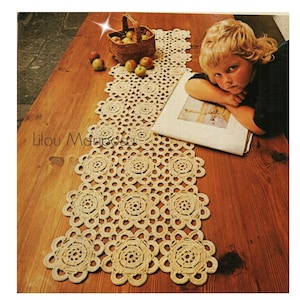 May include: A crocheted table runner with a delicate floral pattern in a cream color. The runner is laid out on a wooden table with a basket of apples and a book.