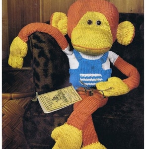 May include: A knitted monkey toy wearing blue overalls with white stripes. The monkey is sitting on a brown chair with a book and glasses in its lap.