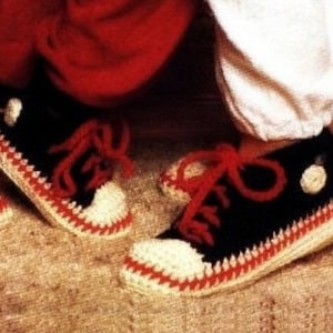 Puede incluir: Un par de zapatillas de crochet negras con cordones rojos y detalles blancos. Las zapatillas tienen una puntera blanca y una franja blanca alrededor de la suela. Las zapatillas están sobre una superficie marrón.