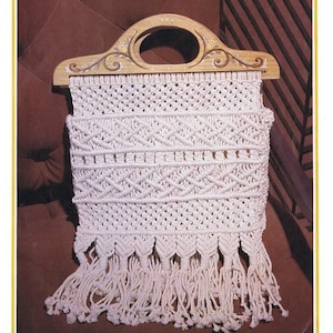 Peut inclure: Un sac en macramé blanc avec une poignée en bois. La poignée est décorée de motifs floraux peints. Le sac a une frange en bas.