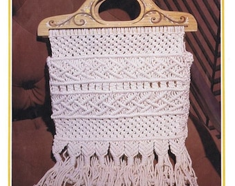 Puede incluir: Un bolso de macramé blanco con un asa de madera. El asa está decorada con diseños florales pintados. El bolso tiene flecos en la parte inferior.