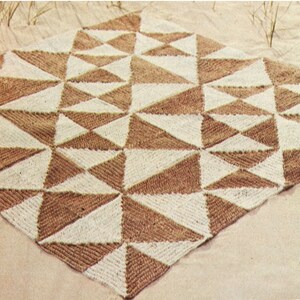 Puede incluir: Una manta tejida con un patrón geométrico de triángulos marrones y crema alternados. La manta es de forma cuadrada y parece estar hecha de un hilo texturizado. El diseño crea un patrón simétrico visualmente atractivo.