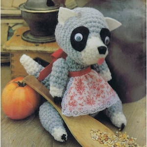 May include: A crocheted raccoon doll wearing a red and white floral apron. The doll is sitting on a wooden spoon with birdseed scattered around it.