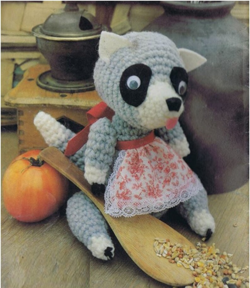 May include: A crocheted raccoon doll wearing a red and white floral apron. The doll is sitting on a wooden spoon with birdseed scattered around it.