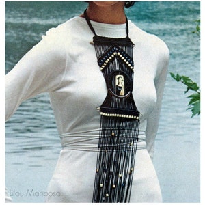 May include: A long, black and white beaded necklace with a circular pendant featuring a woman's face. The necklace is worn by a person wearing a white dress.