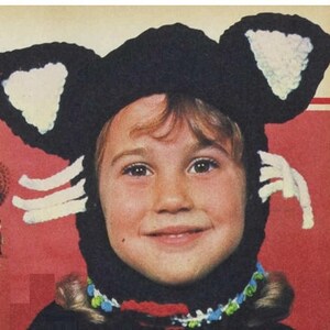 Könnte beinhalten: Ein Kind trägt eine schwarze und weiße gehäkelte Katzenmütze mit weißen Wollohren und Schnurrhaaren. Die Mütze hat ein schwarzes gehäkeltes Gesicht und weiße Wollohren.