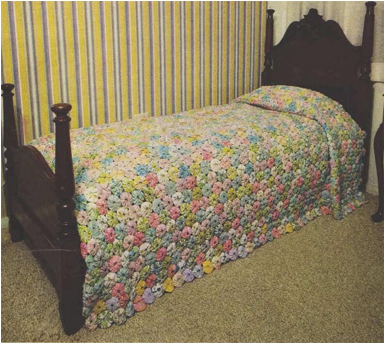 May include: A bed with a wooden headboard and footboard, covered with a colorful crocheted bedspread made of fabric flowers. The bedspread is in shades of pink, blue, yellow, and green.