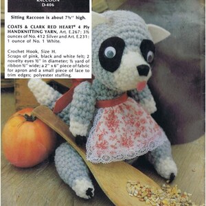 May include: Crocheted raccoon amigurumi toy wearing a pink and white floral dress and a white lace trim. The toy is sitting on a wooden spoon with birdseed scattered around it. The toy is about 7 1/2 inches tall.