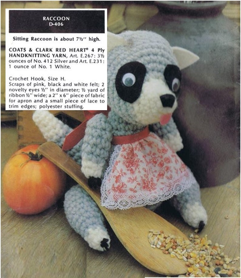 May include: Crocheted raccoon amigurumi toy wearing a pink and white floral dress and a white lace trim. The toy is sitting on a wooden spoon with birdseed scattered around it. The toy is about 7 1/2 inches tall.