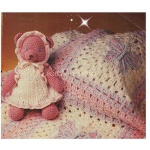 Puede incluir: Una manta de bebé de ganchillo en rosa, blanco y lavanda con un oso de peluche de ganchillo rosa que lleva un vestido blanco. La manta tiene un diseño de mariposa.