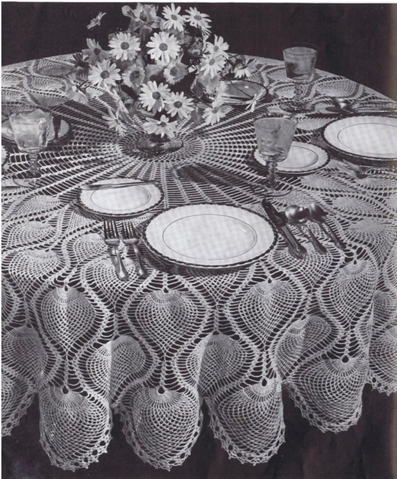 K&ouml;nnte beinhalten: Eine wei&szlig;e H&auml;keltischdecke mit einem zarten, filigranen Muster aus &uuml;berlappenden Kreisen und Muscheln. Die Tischdecke liegt auf einem Tisch mit einer Vase mit wei&szlig;en Blumen in der Mitte und Tellern und Besteck, die darum herum angeordnet sind.