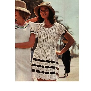 May include: A white crochet dress with a black and white striped band around the bottom. The dress has short sleeves and a round neckline. The model is wearing a straw hat with a white ribbon.