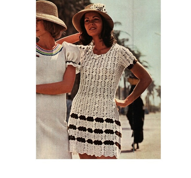 May include: A white crochet dress with a black and white striped band around the bottom. The dress has short sleeves and a round neckline. The model is wearing a straw hat with a white ribbon.