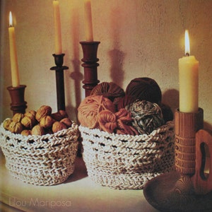 May include: Two crocheted baskets, one filled with walnuts and the other with balls of yarn, sit on a table in front of three lit candles and a wooden candle holder.