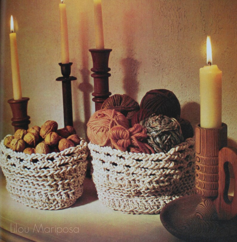 May include: Two crocheted baskets, one filled with walnuts and the other with balls of yarn, sit on a table in front of three lit candles and a wooden candle holder.