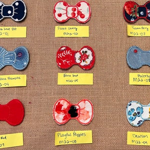 Könnte beinhalten: Neun handgefertigte Schleifen aus Stoff, die auf einem Jute-Hintergrund präsentiert werden. Die Designs umfassen: Julene knot dot, Sweet Cherry, Tuscan Berry, Jolene Fireworks, Jolene knot, Pubrey Chambray, Ruby Red, Playful Poppies und Dawson.