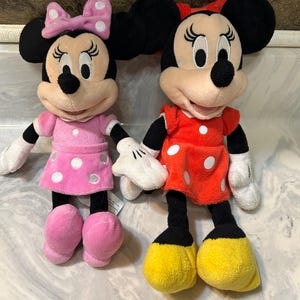 May include: Two plush Minnie Mouse dolls. One doll wears a pink dress with white polka dots and a matching bow. The other doll wears a red dress with white polka dots and a red bow. Both dolls have black ears and black legs.