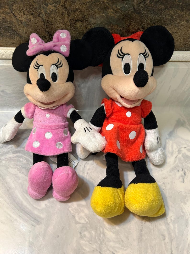 May include: Two plush Minnie Mouse dolls. One doll wears a pink dress with white polka dots and a matching bow. The other doll wears a red dress with white polka dots and a red bow. Both dolls have black ears and black legs.