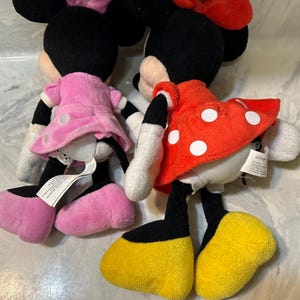 May include: Two Minnie Mouse plush dolls. One doll wears a pink dress and bow, the other a red dress with white polka dots and a red bow. Both have black ears, black legs, white gloves, and yellow shoes.
