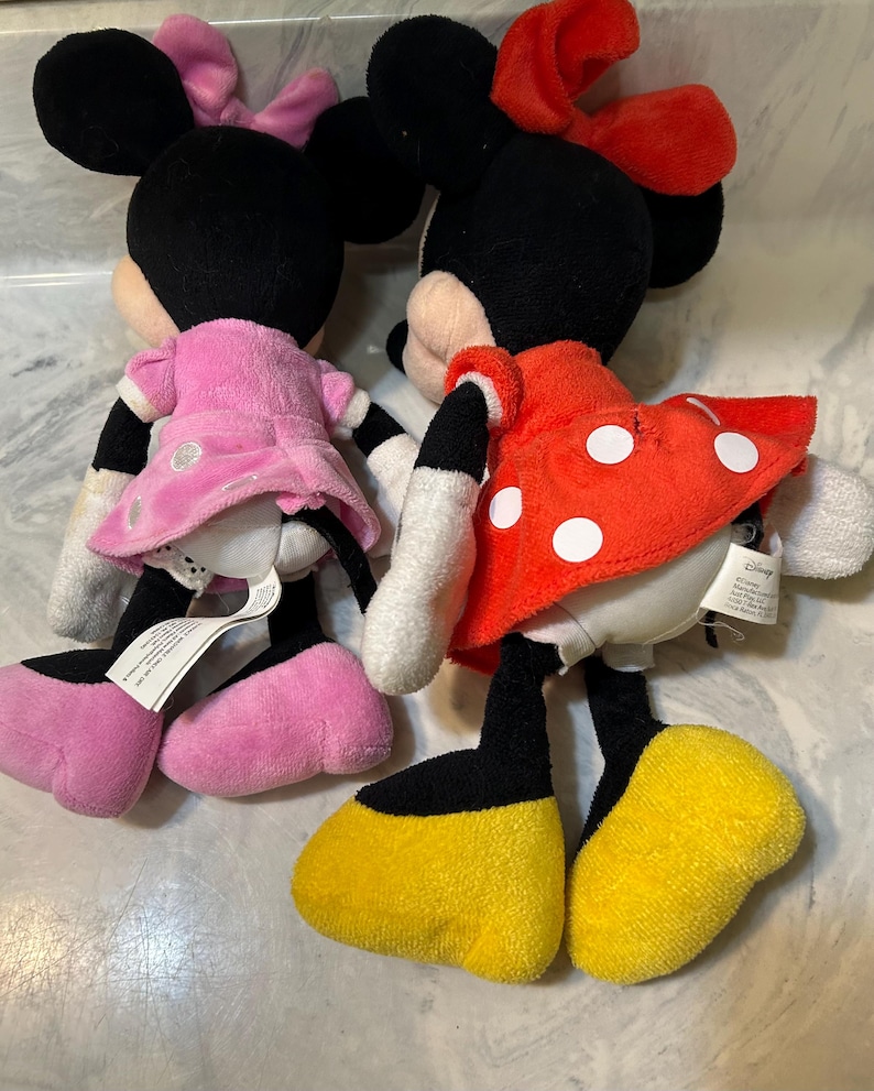 May include: Two Minnie Mouse plush dolls. One doll wears a pink dress and bow, the other a red dress with white polka dots and a red bow. Both have black ears, black legs, white gloves, and yellow shoes.