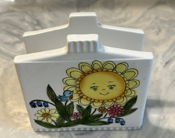 May include: White ceramic napkin holder with a cheerful yellow sunflower design. The sunflower has a smiling face and blue bird perched on a branch. The design also includes green leaves, pink and white flowers, and blue berries.