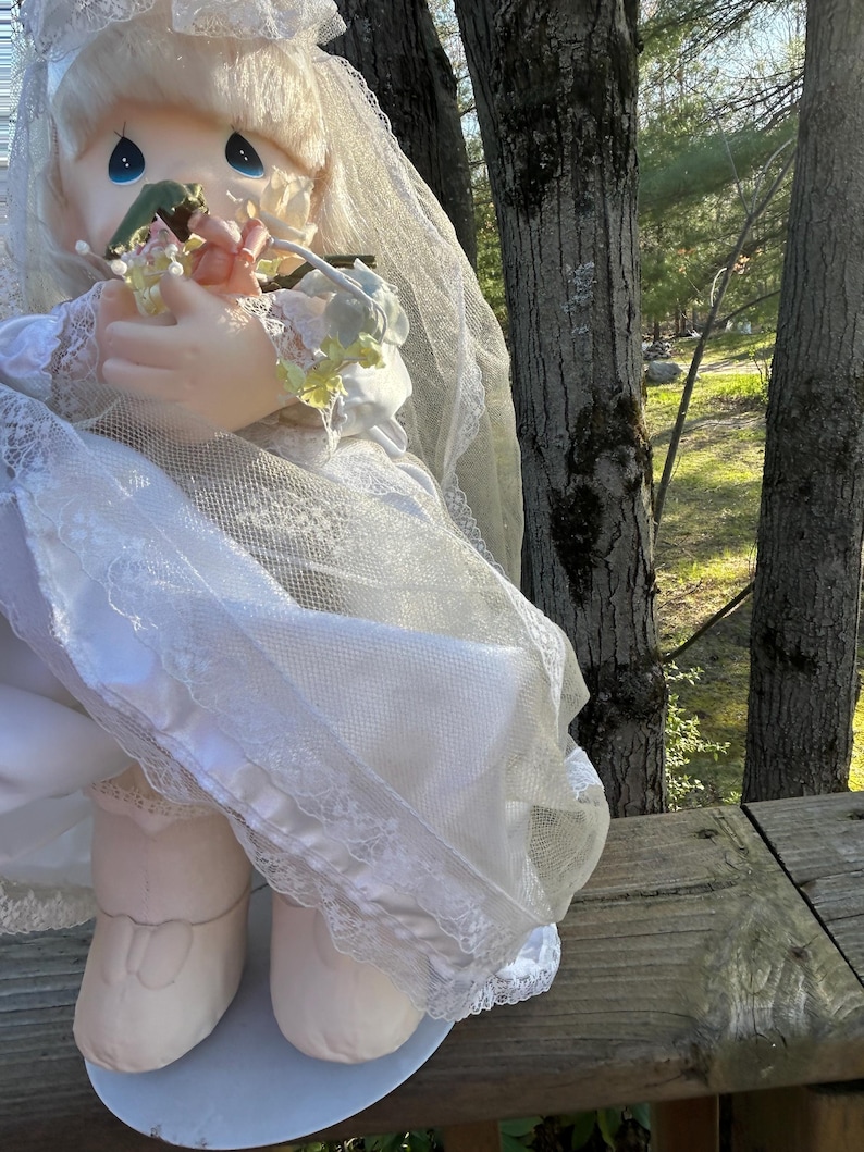Może przedstawiać: Zabytkowa lalka Precious Moments ubrana jako panna młoda, w białym koronkowym welonie i trzymająca mały bukiet kwiat&oacute;w. Lalka ma blond włosy i niebieskie oczy, a także białą satynową suknię z koronkowym wykończeniem.