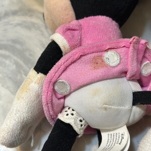 May include: A plush Minnie Mouse doll, featuring a pink dress with white polka dots and a white collar. The doll has black legs and arms, and a white glove. The doll's face is not visible.