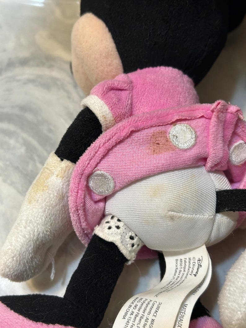 May include: A plush Minnie Mouse doll, featuring a pink dress with white polka dots and a white collar. The doll has black legs and arms, and a white glove. The doll's face is not visible.