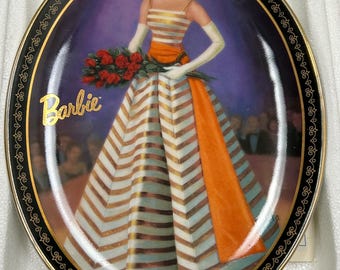 May include: A porcelain plate with a black border and gold trim features a painting of a Barbie doll wearing a white and gold striped dress with an orange sash. The doll is holding a bouquet of red roses and is standing on a stage with an audience in the background. The plate is inscribed with the word "Barbie".