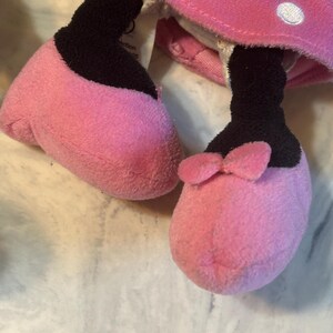 May include: Close-up of a plush toy's feet and lower legs. The feet are pink with black accents and a small pink bow on one foot. The toy's pink dress with white polka dots is visible.