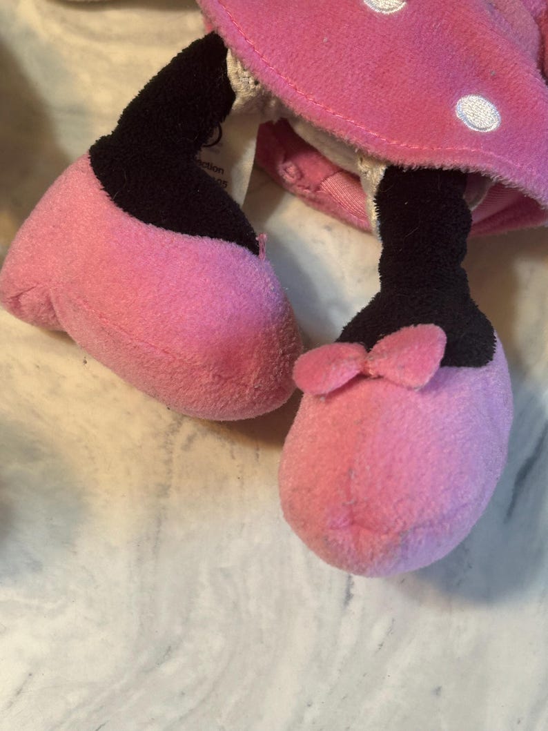 May include: Close-up of a plush toy's feet and lower legs. The feet are pink with black accents and a small pink bow on one foot. The toy's pink dress with white polka dots is visible.