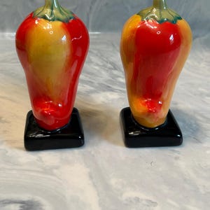 May include: A pair of ceramic salt and pepper shakers shaped like chili peppers. Each pepper features a glossy gradient of red and yellow with a green stem at the top. The shakers are mounted on square, solid black bases for stability.